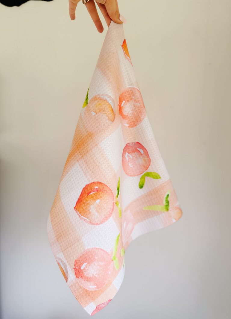 A hand holds up a patterned cloth featuring a design of red apples and green leaves on a white and beige checkered background. - Spruced Studio Upscale Workshop