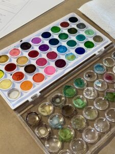 A watercolor paint palette with multiple colors, some of which have been used; a sheet of white paper towel sits nearby on a brown surface. - Spruced Studio Upscale Workshop