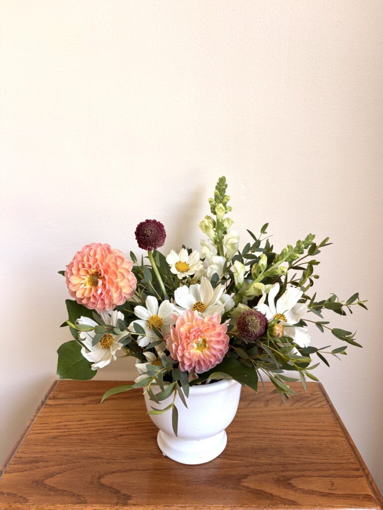 A white vase with a floral arrangement of pink dahlias, white cosmos, snapdragons, and greenery sits on a wooden table against a plain beige wall. - Spruced Studio Upscale Workshop