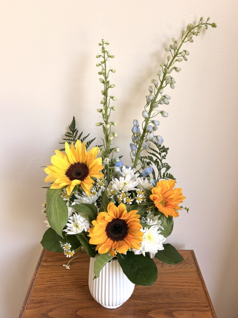 A white vase with sunflowers, white and blue flowers, green leaves, and tall flower spikes sits on a wooden table against a light-colored wall. - Spruced Studio Upscale Workshop