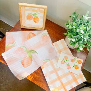 A wooden table with a potted plant, peach-themed towel, art print, notecard, and a framed peach illustration arranged neatly. - Spruced Studio Upscale Workshop