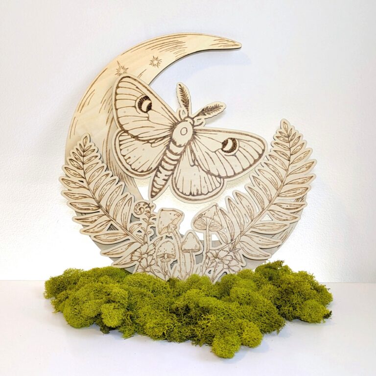 Wooden artwork featuring a moth, crescent moon, ferns, and mushrooms, displayed with a base of green moss against a white background. - Spruced Studio Upscale Workshop