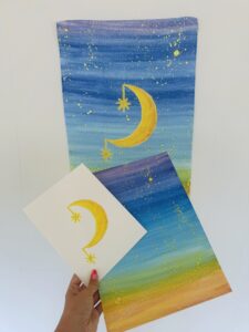 A hand holds two artworks with yellow crescent moons and stars on blue, green, and yellow gradient backgrounds against a white wall. - Spruced Studio Upscale Workshop