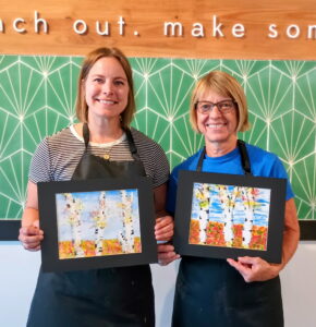 Two women wearing aprons stand indoors, each holding a framed birch tree painting. A green geometric wall and partial lettering are visible in the background. - Spruced Studio Upscale Workshop