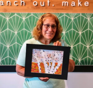 A person wearing glasses holds up a framed artwork of birch trees with autumn foliage, standing in front of a green geometric-patterned wall. - Spruced Studio Upscale Workshop