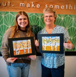Two women standing side by side indoors, each holding a framed painting of birch trees with autumn foliage. A green patterned wall and motivational sign are visible in the background. - Spruced Studio Upscale Workshop
