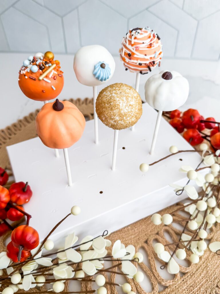 Six decorated cake pops on white sticks stand upright in a foam block, surrounded by artificial berries and autumn-themed decorations on a woven mat. - Spruced Studio Upscale Workshop