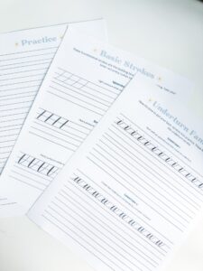 Three sheets of calligraphy practice worksheets showing practice, basic strokes, and underturn fan drills, with lined guides and sample letters on a white surface. - Spruced Studio Upscale Workshop