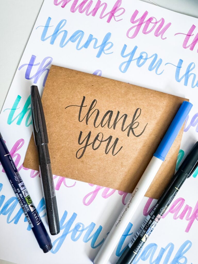 A brown envelope with “thank you” written on it is placed on a paper with repeated “thank you” text, surrounded by pens and markers. - Spruced Studio Upscale Workshop