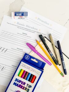 A hand lettering practice sheet with pens, markers, a pencil, an eraser, and a case arranged on a white surface. - Spruced Studio Upscale Workshop