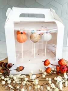 Four decorative cake pops, shaped and colored like pumpkins and fall themes, displayed inside a white box with a clear plastic window. Autumn-themed artificial berries and leaves in front. - Spruced Studio Upscale Workshop