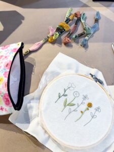 Embroidery hoop with floral designs on white fabric, colored threads, and a floral zipper pouch on a beige surface. - Spruced Studio Upscale Workshop