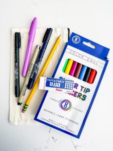 A set of pens, a yellow pencil, an eraser, and a pack of colorful markers are displayed on a white surface with a small cloth pouch. - Spruced Studio Upscale Workshop