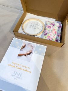 Beginner’s hand embroidery kit with instruction booklet, hoop with fabric, and floral cloth, displayed inside an open cardboard box on a table. - Spruced Studio Upscale Workshop