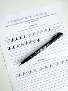 A worksheet titled "Underturn Family" with calligraphy practice for lowercase "i" and "u" and a black pen resting on the page. - Spruced Studio Upscale Workshop
