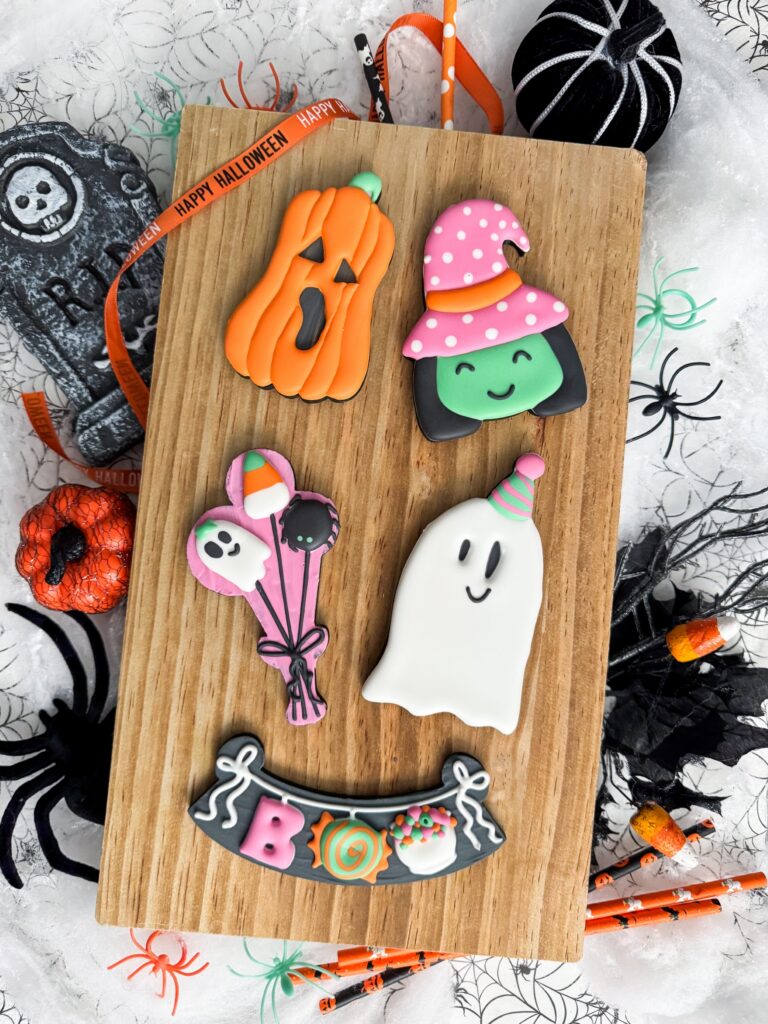Five decorated Halloween cookies—pumpkin, witch, ghost, balloon bunch, and “BOO” banner—on a wooden board, surrounded by festive decorations. - Spruced Studio Upscale Workshop