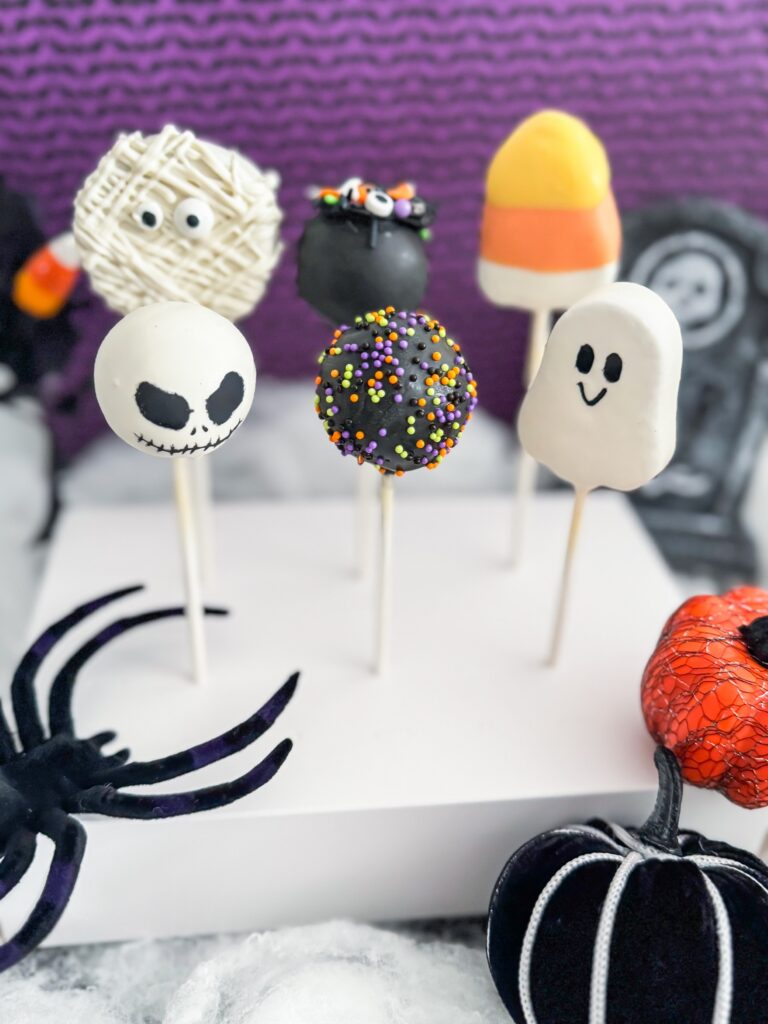 Six Halloween-themed cake pops are displayed upright, decorated as a mummy, monster, candy corn, Jack Skellington, ghost, and a sprinkle-covered pop; Halloween props surround them. - Spruced Studio Upscale Workshop