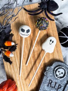 Three Halloween cake pops—a skull, a ghost, and a sprinkle-covered pop—on sticks, displayed on a wooden board with themed decorations like candy corn, a spider, and a tombstone. - Spruced Studio Upscale Workshop