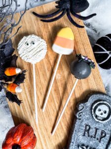 Three Halloween-themed cake pops on sticks—a mummy, a candy corn, and a black cauldron—on a wooden board with Halloween decorations, including a spider, candy, and tombstone. - Spruced Studio Upscale Workshop