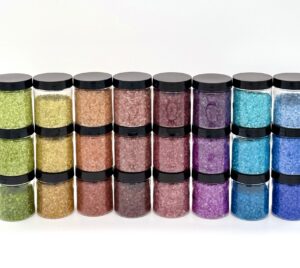 Five rows of clear plastic jars with black lids, each filled with different colors of decorative crushed glass, arranged in a grid pattern against a plain white background. - Spruced Studio Upscale Workshop