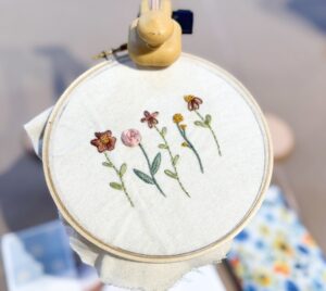 An embroidery hoop holds fabric with hand-stitched flowers, surrounded by blurred objects including a floral pouch and a booklet. - Spruced Studio Upscale Workshop