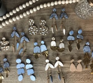 A collection of handmade polymer clay earrings in blue, white, and tan tones with various shapes and patterns displayed on a wooden surface. - Spruced Studio Upscale Workshop