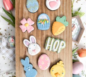 Nine decorated Easter-themed sugar cookies, including eggs, a bunny, a chick, a carrot, a basket, a flower, and the word "HOP," arranged on a wooden board with spring decorations surrounding them. - Spruced Studio Upscale Workshop