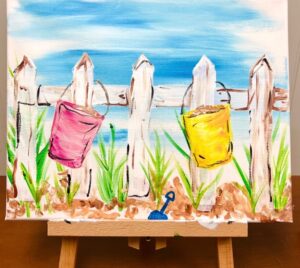 A painting on an easel depicts a white fence with a pink and a yellow bucket hanging on it, green grass, brown sand, and a blue sky in the background. - Spruced Studio Upscale Workshop