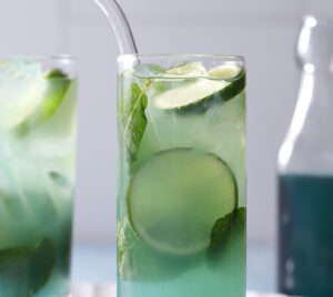 A tall glass of blue-green iced drink with lime slices, mint leaves, and a clear straw, set on a light surface with another similar drink and a bottle in the background. - Spruced Studio Upscale Workshop