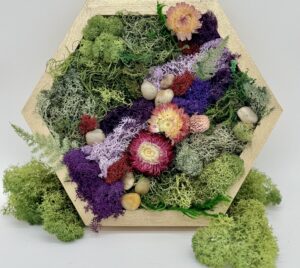 A hexagonal frame displays preserved moss, purple florets, dried flowers, and small decorative stones, arranged artistically against a plain white background. - Spruced Studio Upscale Workshop